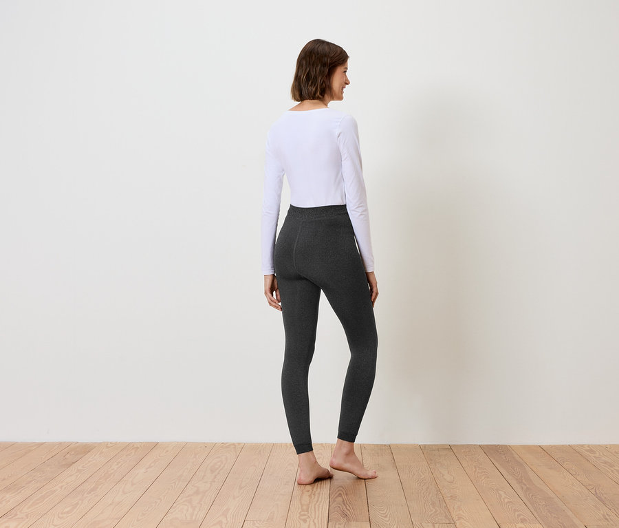 Eine Frau steht mit dem Rücken zur Kamera und trägt ein weißes Langarmshirt und graue Leggings.