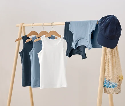 Auf einem hölzernen Kleiderständer hängen drei Tanktops auf Bügeln, daneben liegen zwei weitere Tanktops, eine blaue Schirmmütze und eine Netztasche.