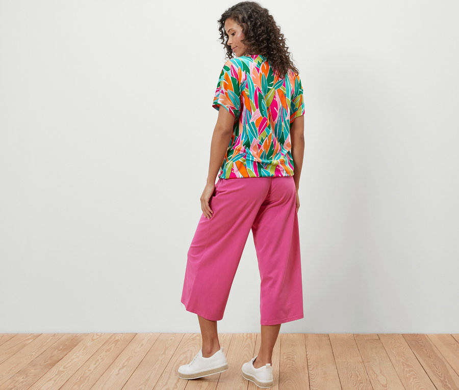 Frau von hinten in überschnittenem Blusenshirt mit Alloverprint und pinker Hose.