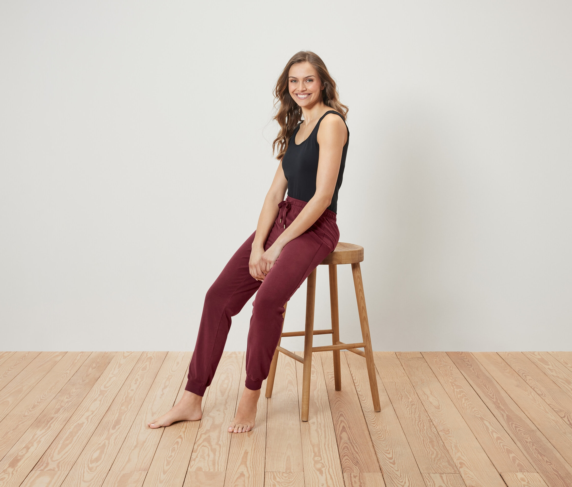 Lächelnde Frau sitzt auf einem Holzhocker und trägt ein schwarzes Tanktop und bordeauxrote Jogginghose.