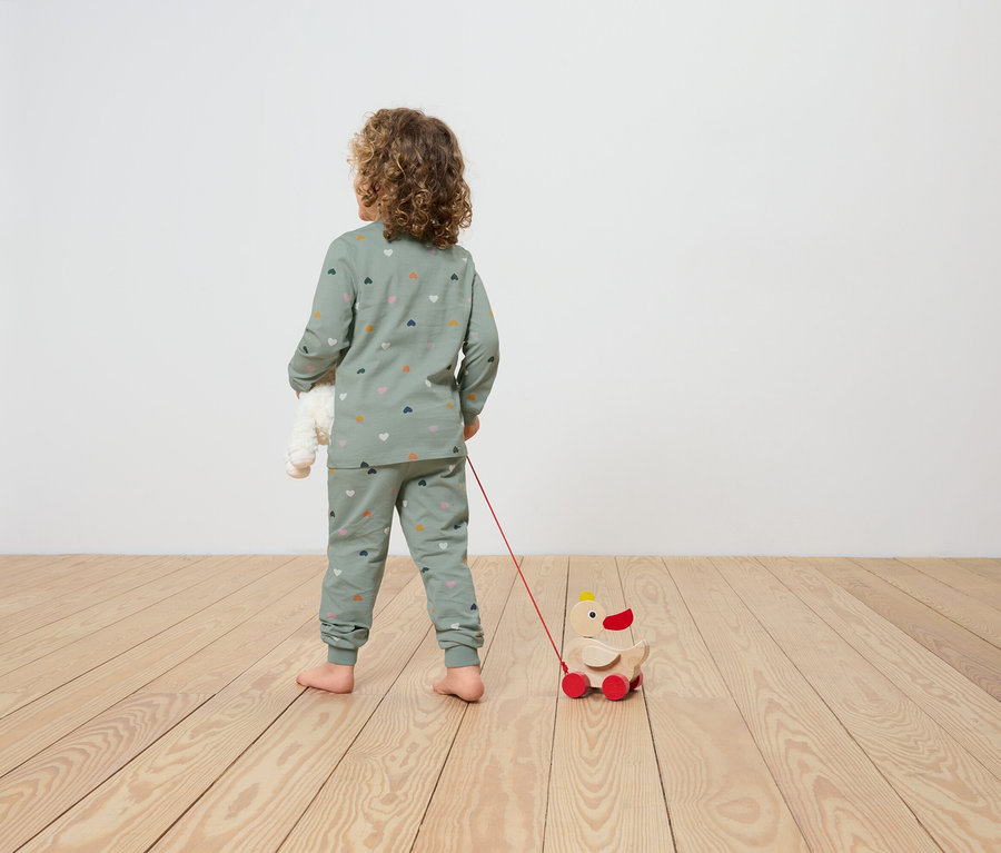 Ein Kind im Schlafanzug mit Herzprint zieht ein hölzernes Entenspielzeug auf Rädern hinter sich her und hält einen Teddybären im Arm.