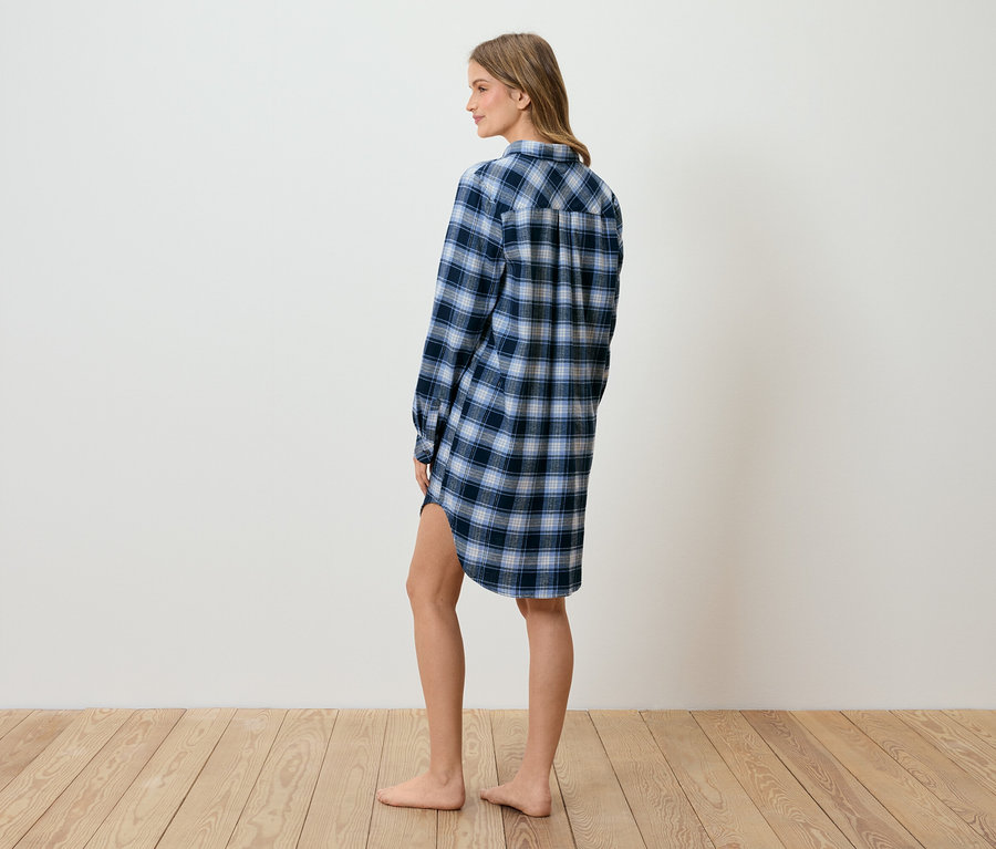 Frau im blau karierten Hemdkleid steht seitlich auf einem Holzboden vor einer weißen Wand.
