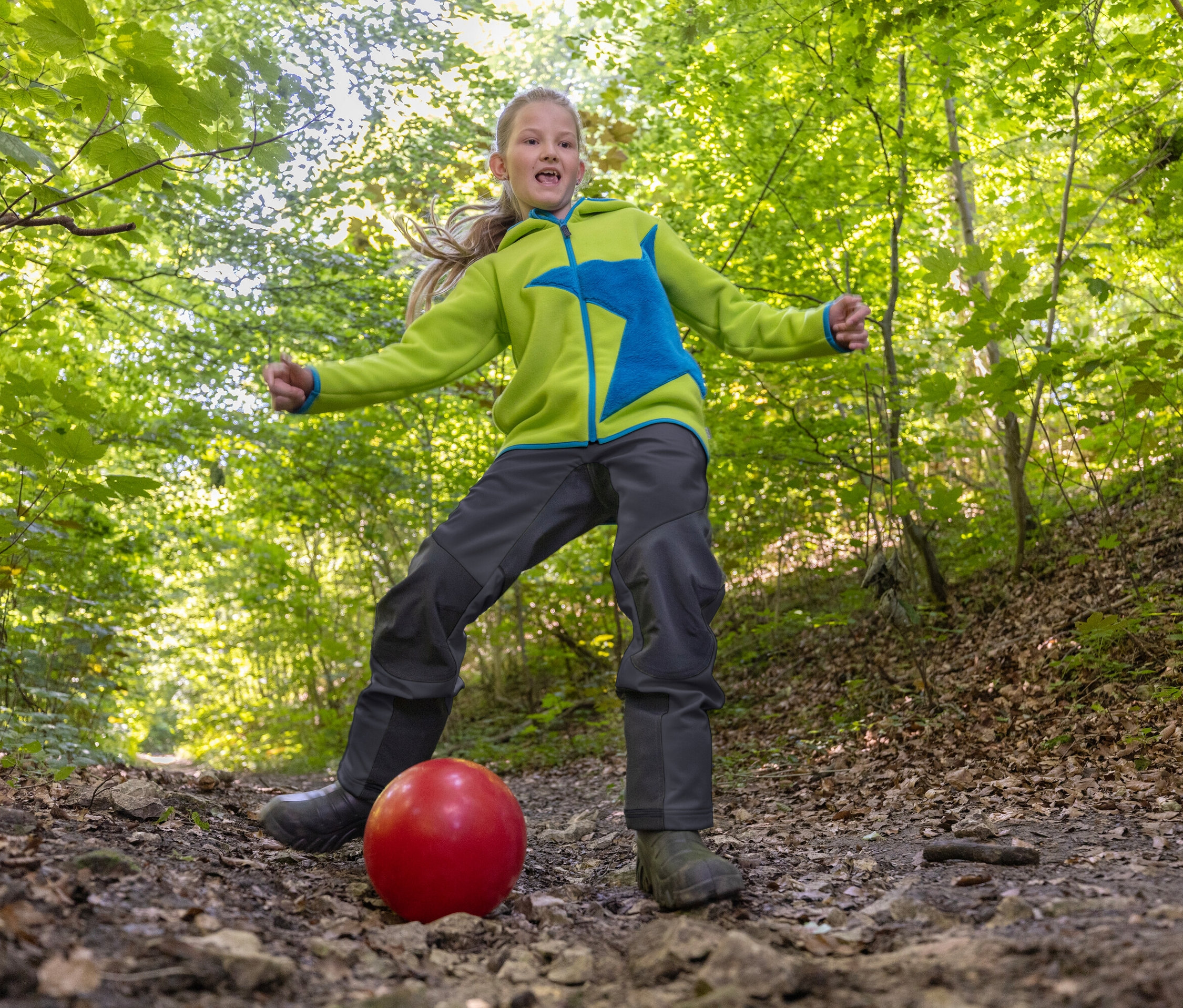 Ein Mädchen tritt einen roten Ball auf einem Waldweg. Das Mädchen trägt eine grüne Jacke mit einem blauen Stern und dunkle Hosen.