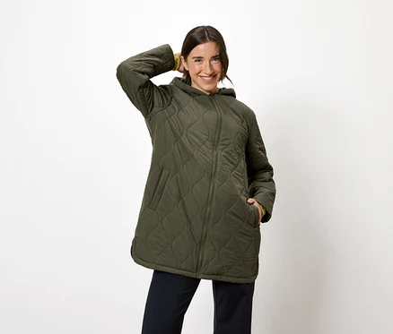 Frau posiert in olivgrüner Steppjacke mit Kapuze und marineblauer Hose.