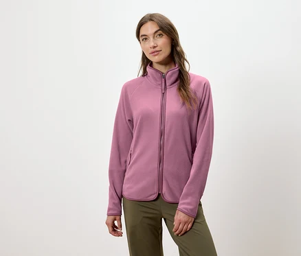 Eine Frau trägt eine rosafarbene Fleecejacke mit Reißverschluss und eine grüne Hose.