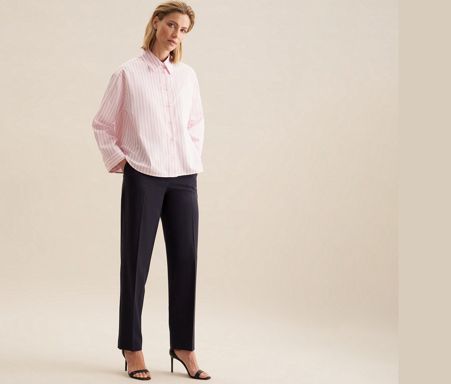 Eine Frau trägt eine Seidensticker Oversize Hemdbluse in Rosé, dazu dunkle Hosen und schwarze Sandaletten mit Absatz.