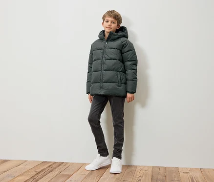 Ein Junge steht und trägt eine dunkelgrüne Daunenjacke, graue Jeans und weiße Turnschuhe.