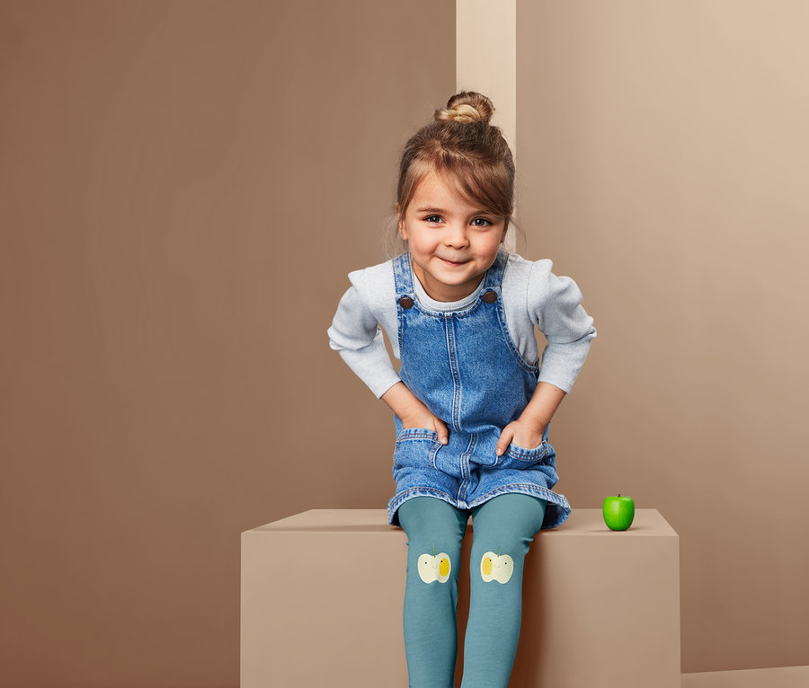 Ein Mädchen sitzt und trägt ein Jeanskleid und 2 Leggings mit Flicken.