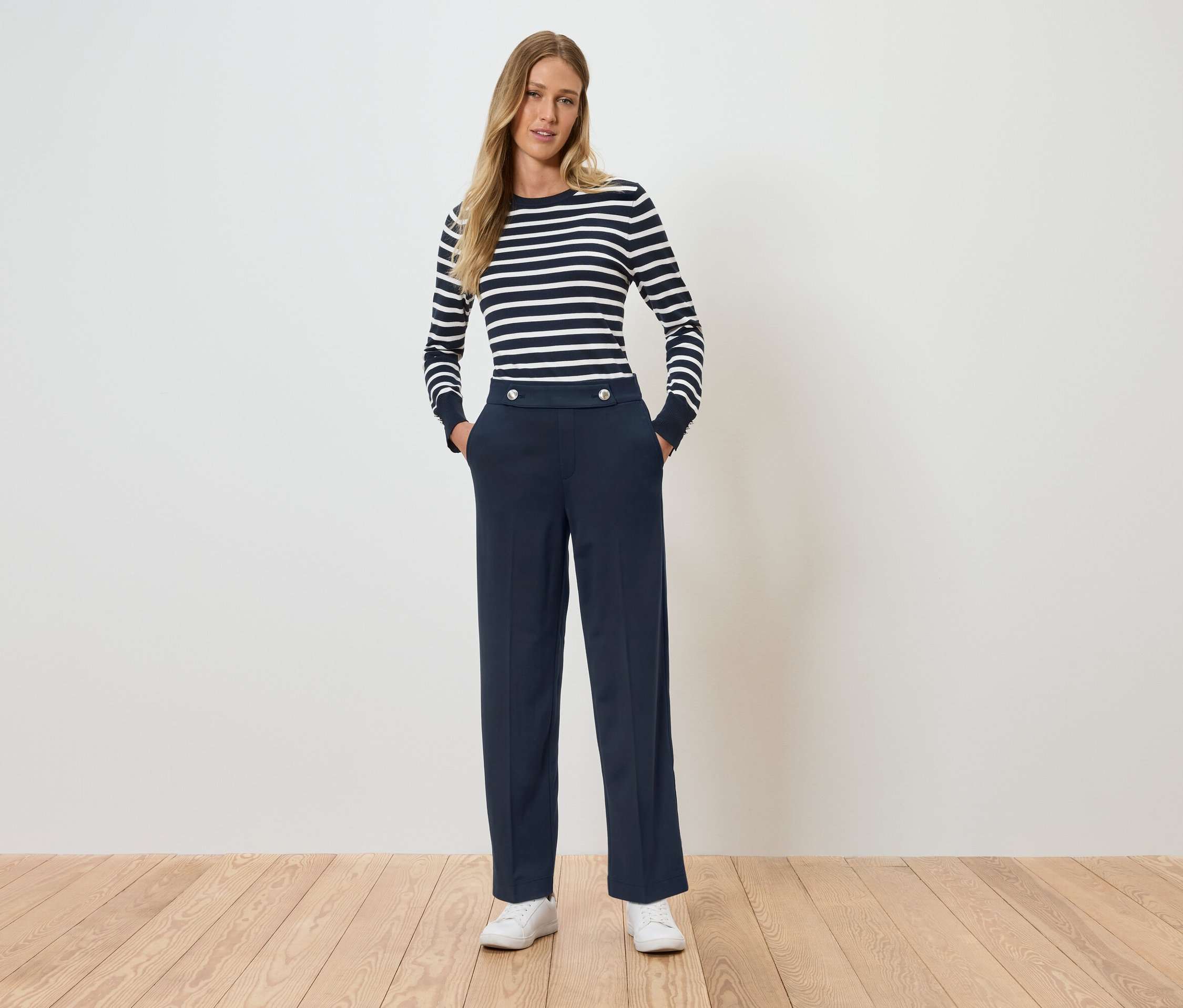 Eine Frau steht mit den Händen in den Hosentaschen. Sie trägt ein gestreiftes Langarmshirt und dunkelblaue Hosen.