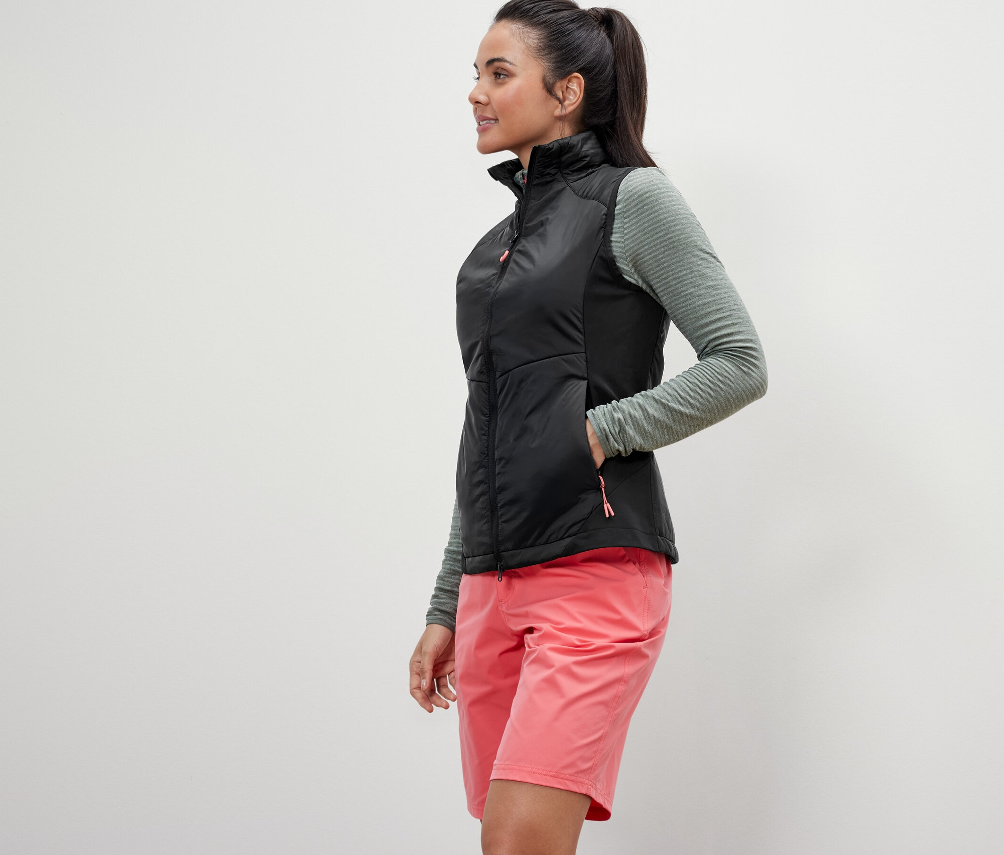 Frau posiert in Hybrid-Weste »pack light«, olivgrünem Oberteil und pinkfarbenen Shorts.