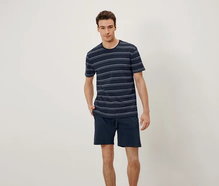 Ein Mann steht und hat seine Hände in den Hosentaschen. Er trägt ein T-Shirt mit horizontalen Streifen und Shorts.
