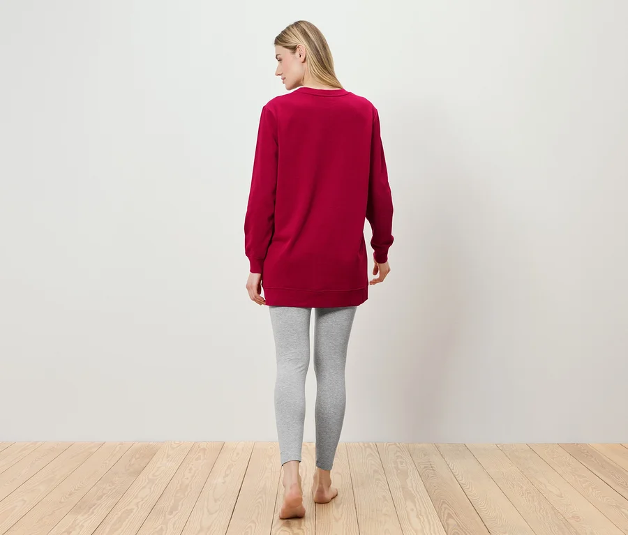 Eine Frau steht mit dem Rücken zur Kamera und trägt eine weinrote Tunika und graue Leggings.