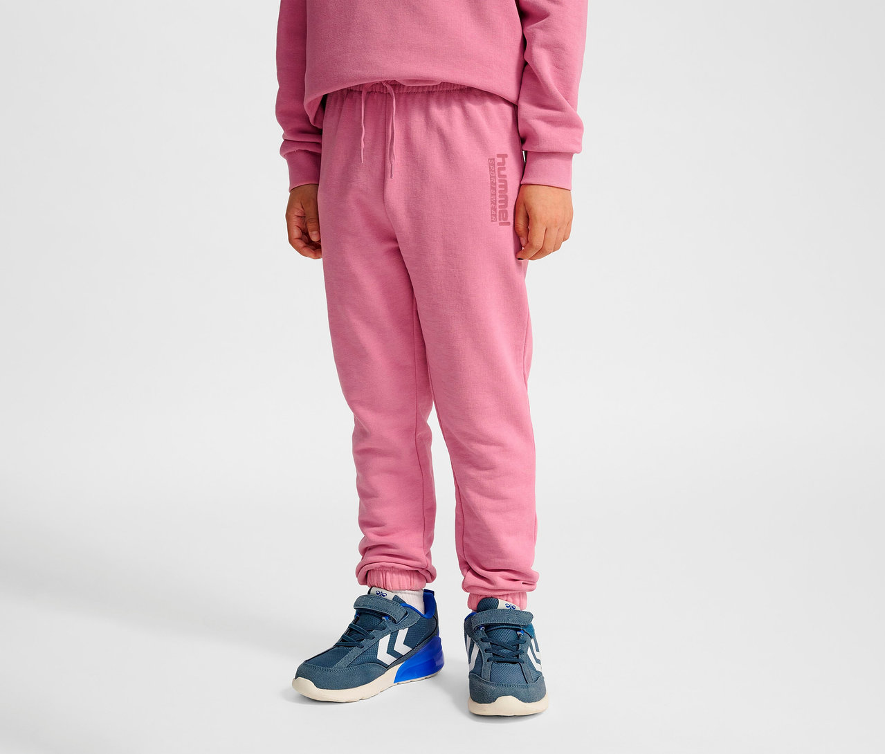 Junge trägt eine rosafarbene HUMMEL HMLJR Base Regular Pants und blaue Turnschuhe.