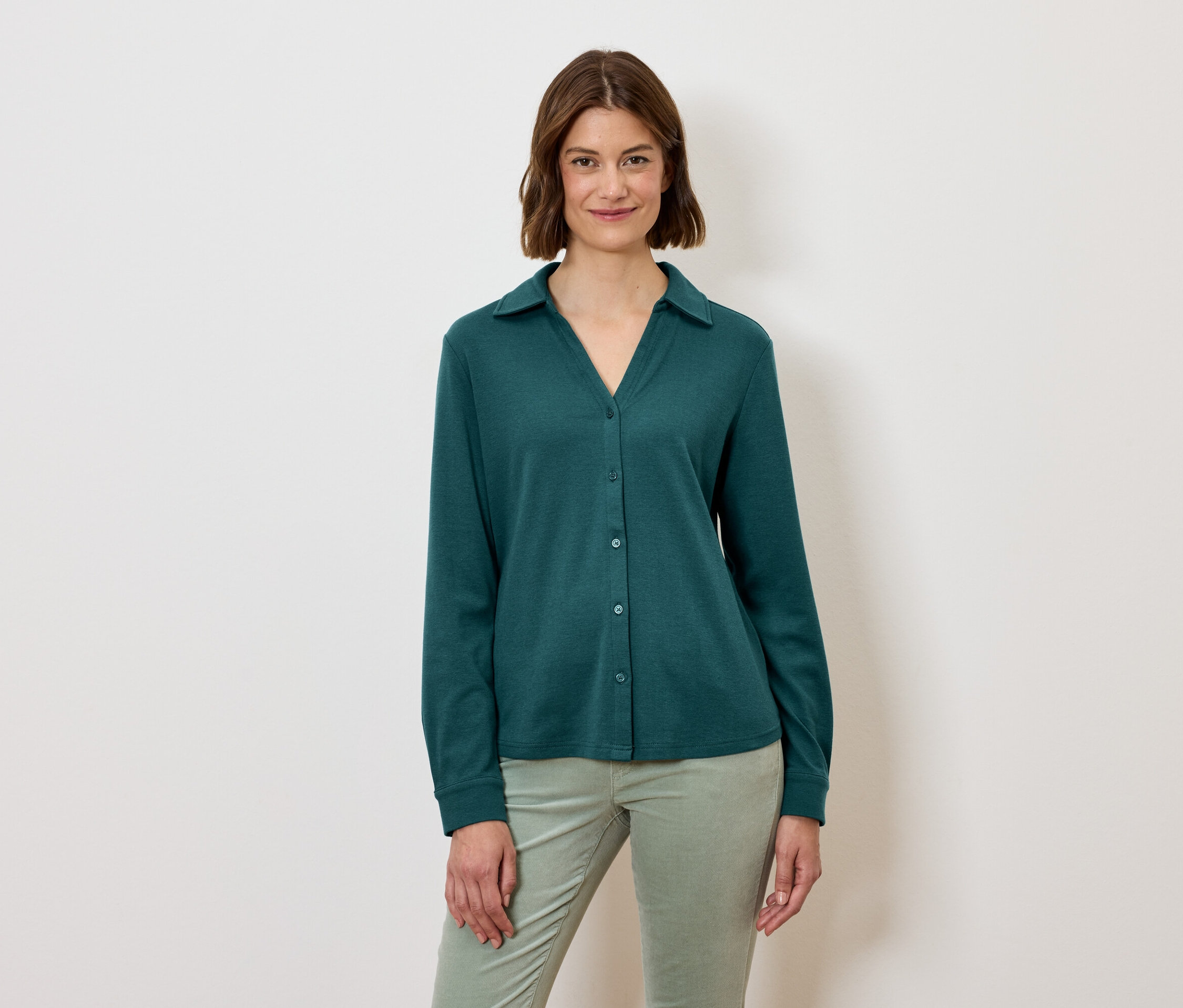 Frau mit kurzem braunem Haar trägt ein grünes, langärmeliges Hemd und hellgrüne Hosen.