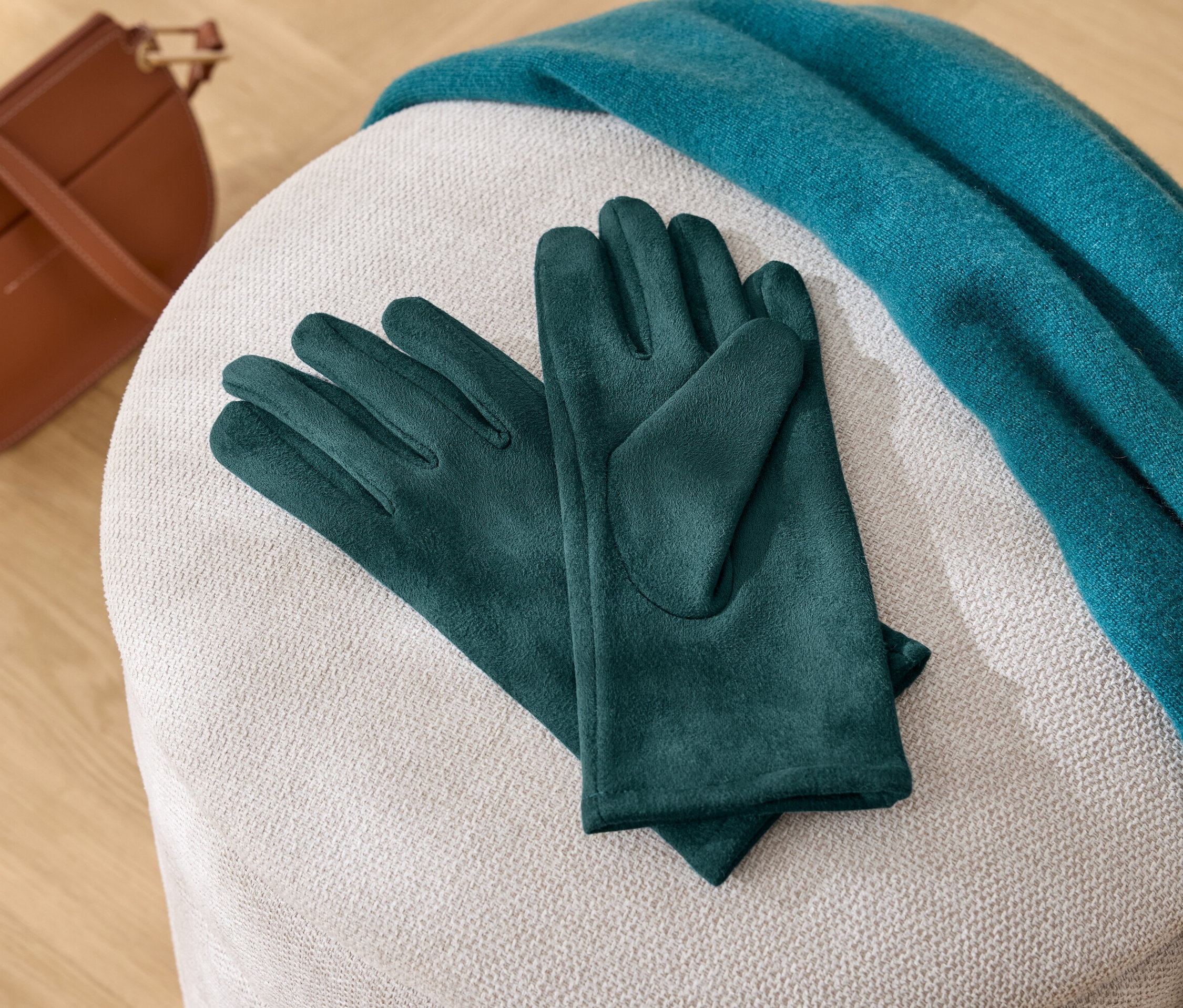 Auf einem weißen Hocker liegen grüne Handschuhe, ein blauer Schal und eine braune Handtasche.