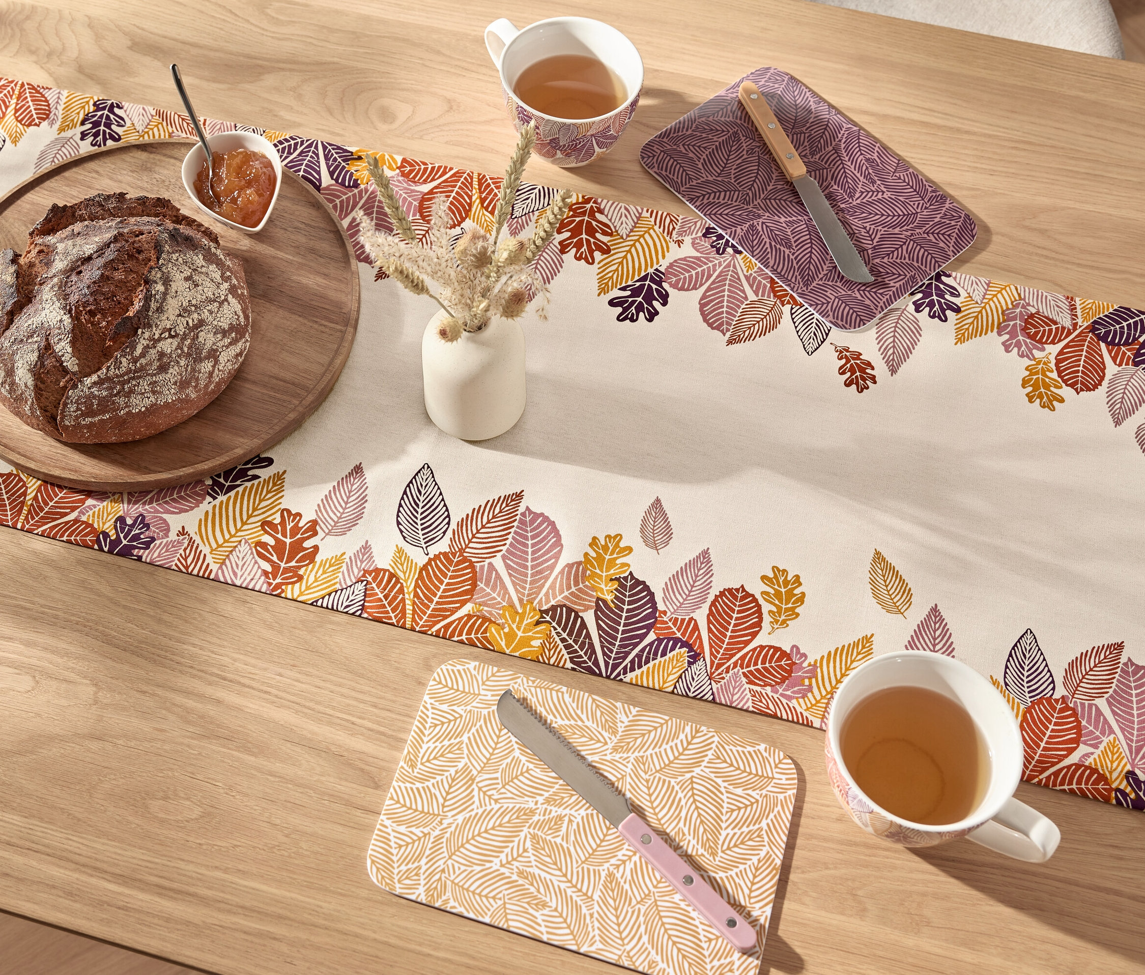 Brot auf Holzbrett und eine Tasse Tee auf einem Tischläufer mit Blättermuster.