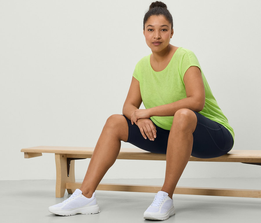 Frau sitzt auf Bank in einem Sportshirt, lime und Sportradler, dunkelblau.