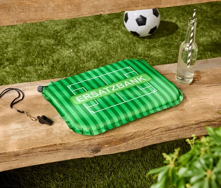 Auf einer Holzbank liegen ein grünes Kissen mit der Aufschrift 'ERSATZBANK', eine Pfeife, eine Wasserflasche und ein Fußball.