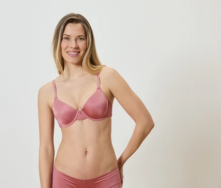 Frau mit blonden Haaren, die einen rosafarbenen BH und passende Unterwäsche vor einem weißen Hintergrund trägt.