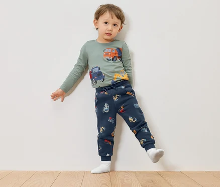 Ein Kind mit einem grünen Langarmshirt mit Bauwagenmotiven und marineblauen Hosen mit Bauwagenmotiven steht vor einer weißen Wand.