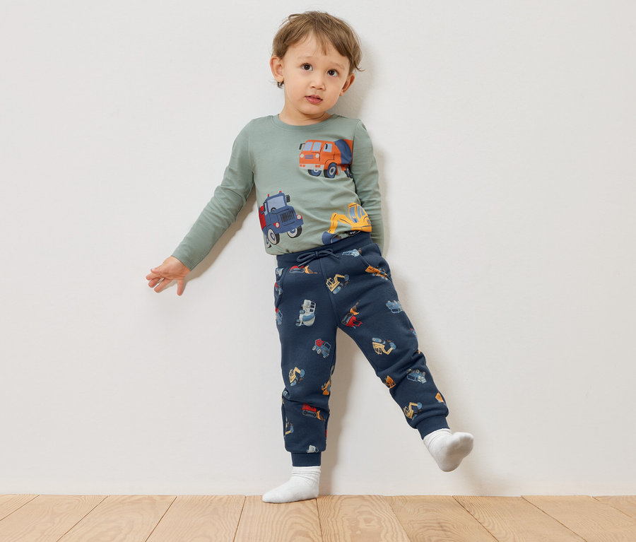Ein Kind mit einem grünen Langarmshirt mit Bauwagenmotiven und marineblauen Hosen mit Bauwagenmotiven steht vor einer weißen Wand.