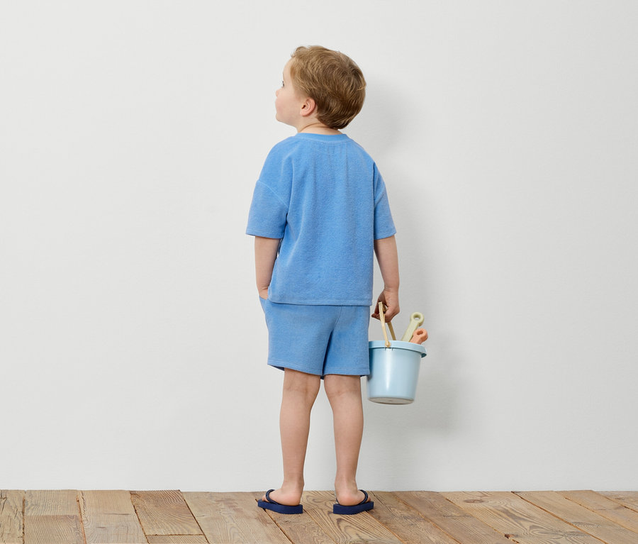 Junge im blauen Frottee-Set hält blauen Eimer mit Spielzeug vor weisser Wand.