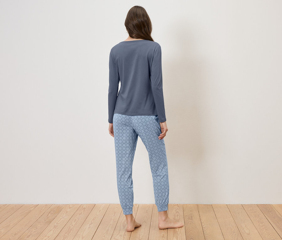 Eine Frau von hinten in einem blauen Schlafanzug mit Muster, stehend auf einem Holzboden vor einer weißen Wand.