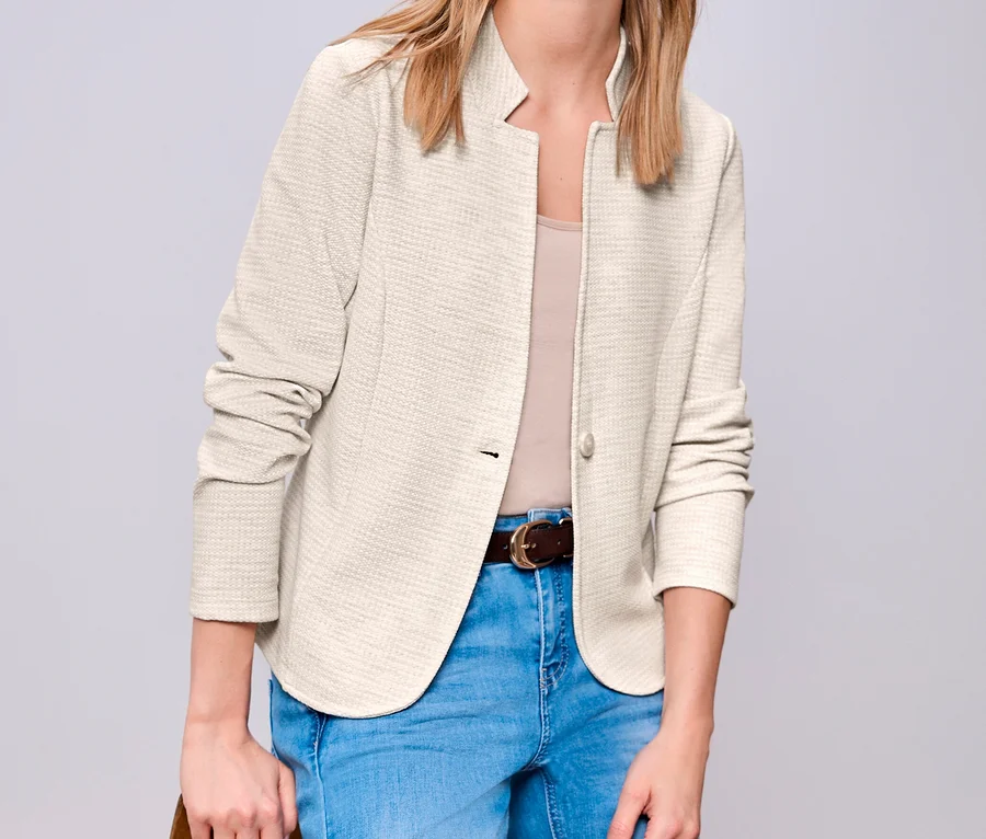 Blonde Frau trägt einen beigefarbenen Street One Jerseyblazer, hellblaue Jeans und einen braunen Gürtel, steht mit Hand auf Holzstuhl.