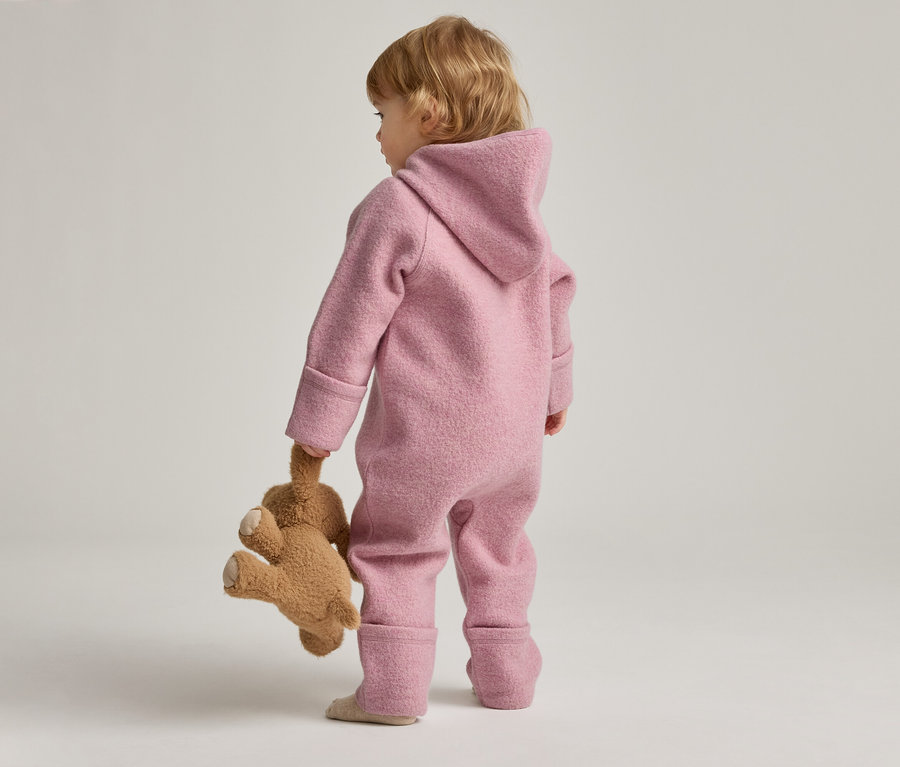 Kleinkind in rosafarbenem Overall mit Kapuze hält Teddybär in der Hand, von hinten gesehen.