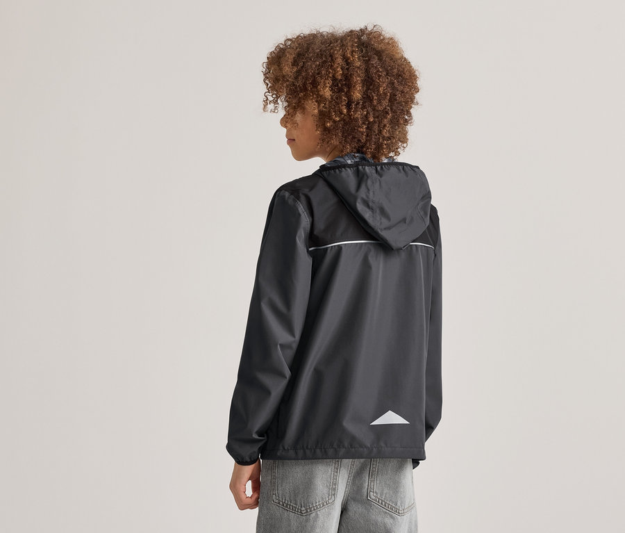 Person mit lockigem Haar von hinten, trägt eine schwarze Regenjacke mit Kapuze und reflektierenden Details, dazu graue Jeans.