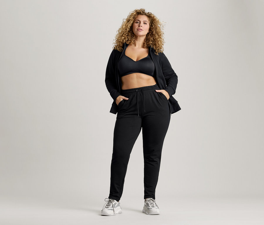 Frau mit lockigem Haar, die einen schwarzen Sport-BH, eine Jacke und eine Jogginghose trägt und die Hände in den Taschen hat.