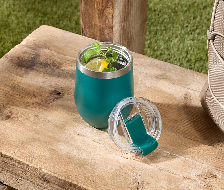 Ein grüner, isolierter Becher mit einem Getränk mit Zitrone und Minze steht mit seinem Deckel auf einem Holztisch neben einer beigen Tasche.