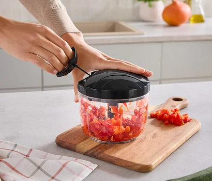 Hände bedienen einen manuellen Zerkleinerer mit gehacktem rotem Gemüse. Der Zerkleinerer steht auf einem Holzschneidebrett mit weiteren Gemüsestücken. Ein kariertes Küchentuch und eine Zucchini liegen auf der Arbeitsplatte.