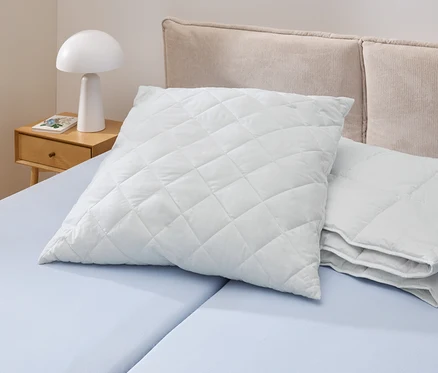 Ein weißes, gestepptes Kopfkissen Cell Solution® (90 x 70 cm) liegt auf einem hellblauen Bett. Daneben liegen weitere Kopfkissen Cell Solution® (80 x 80 cm, 100 x 65 cm).