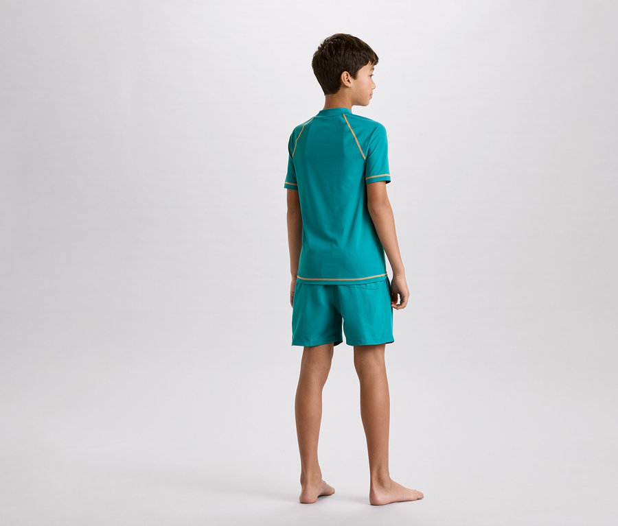 Junge von hinten, trägt türkisfarbenes T-Shirt und Shorts mit goldenen Ziernähten.