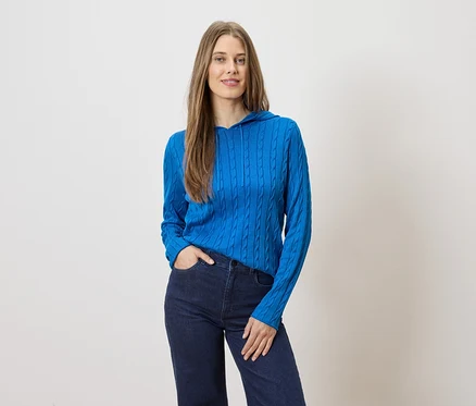 Frau mit langen Haaren posiert in blauer Strickjacke mit Kapuze und dunkler Jeans vor hellem Hintergrund.