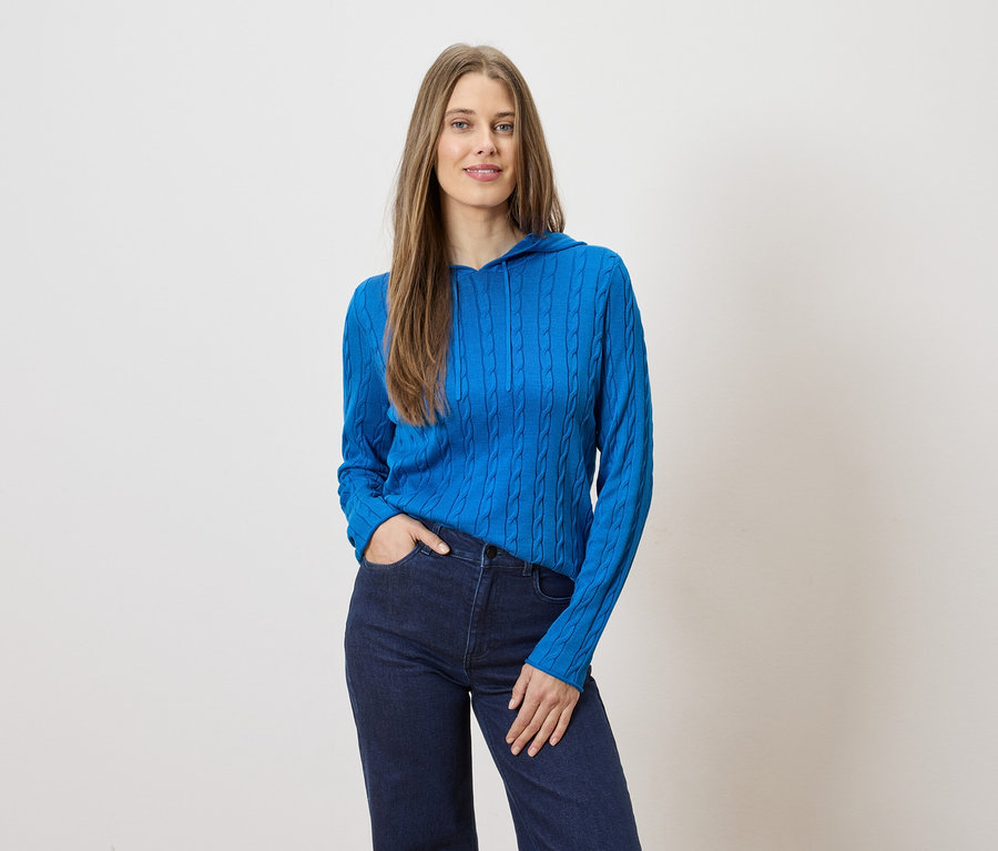 Frau mit langen Haaren posiert in blauer Strickjacke mit Kapuze und dunkler Jeans vor hellem Hintergrund.