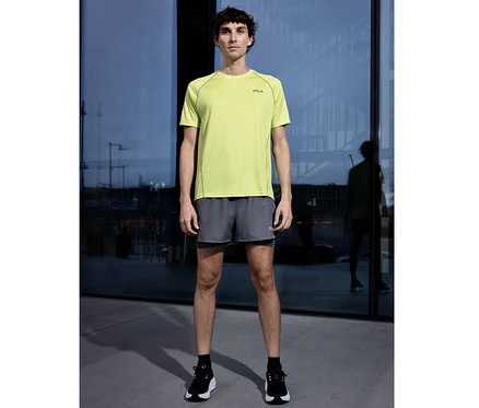 Mann steht vor Glasfassade, trägt ein hellgrünes FILA Herren-Funktionsshirt, graue Shorts und schwarze Sportschuhe.