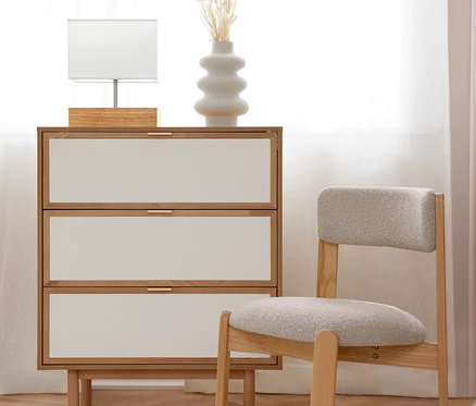 Eine helle Holzkommode mit drei weißen Schubladen und goldenen Griffen, darauf eine weiße Tischlampe und eine weiße Vase mit Trockenblumen. Daneben steht ein heller Holzstuhl mit grau gepolsterter Sitzfläche. Der Raum hat einen Holzboden mit einem weißen Teppich und ein helles Fenster mit weißen Vorhängen.