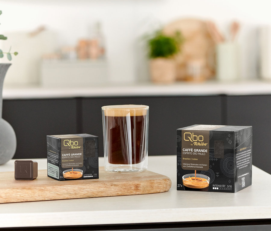 Auf einem Tisch stehen eine Tasse Kaffee und Packungen Qbo Caffè Grande ESPÍRITO SÃO PAULO mit 8 und 27 Kapseln.