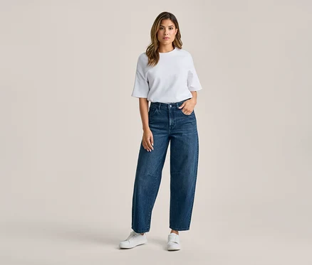 Frau steht, trägt ein weißes T-Shirt, dunkelblaue weite Jeans und weiße Sneaker. Ihre rechte Hand steckt in der Hosentasche.