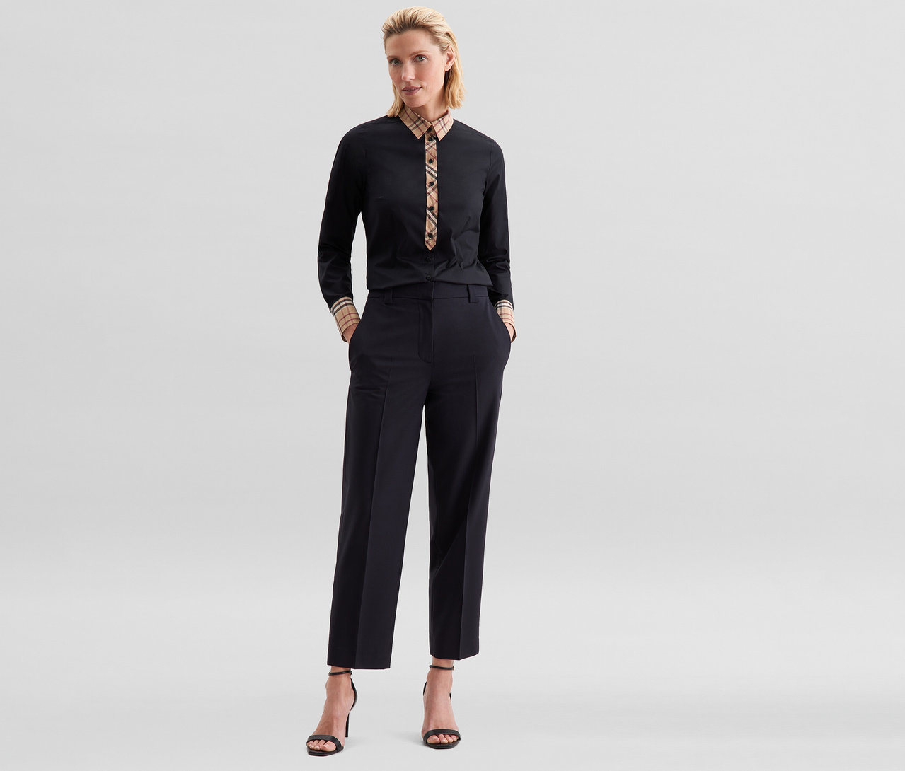 Frau steht mit Händen in den Taschen, trägt eine schwarze Seidensticker Popeline-Bluse mit Karodetail, schwarze Cropped-Hose und schwarze Sandalen mit hohen Absätzen.