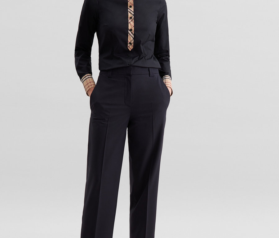 Eine Frau steht mit Händen in den Hosentaschen und trägt eine schwarze Seidensticker Popeline-Bluse mit Karodetail an Kragen und Manschetten sowie eine dunkle Hose und schwarze Sandaletten.