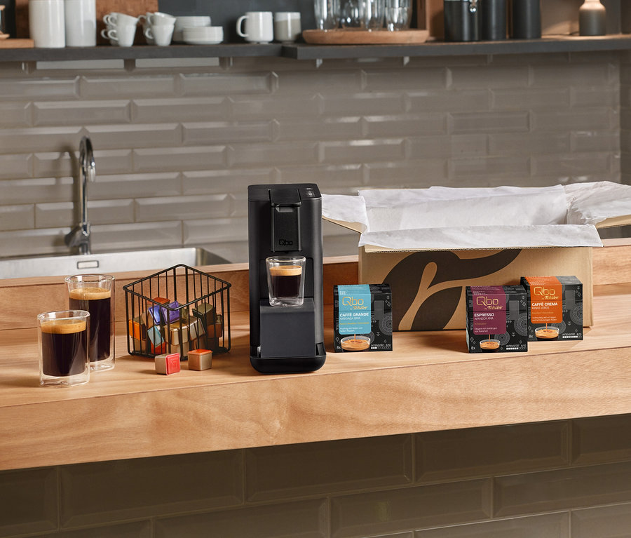 Die Qbo Iconic, Dark Chrome 1,1 DAEE Kaffeemaschine mit einem Glas Kaffee darunter, daneben zwei weitere Gläser Kaffee und ein Korb voller Kapseln. Mehrere Packungen Qbo Espresso APANECA ANA, Qbo Caffè Grande KIRINYAGA BAYA und Qbo Caffè MINAS VERDE Kapseln stehen neben einem offenen Karton auf der Küchentheke.