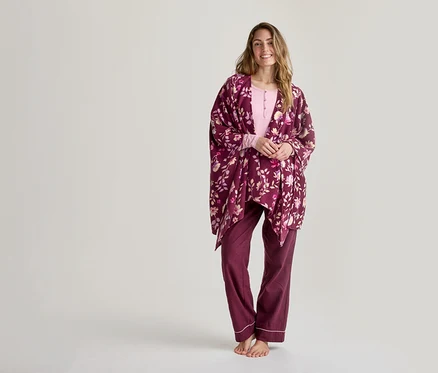 Lächelnde Frau in einem hellrosa Langarmshirt, einer dunkelroten Pyjamahose mit weißer Paspelierung und einem dunkelroten floralen Wickelmantel steht barfuß vor einem weißen Hintergrund.