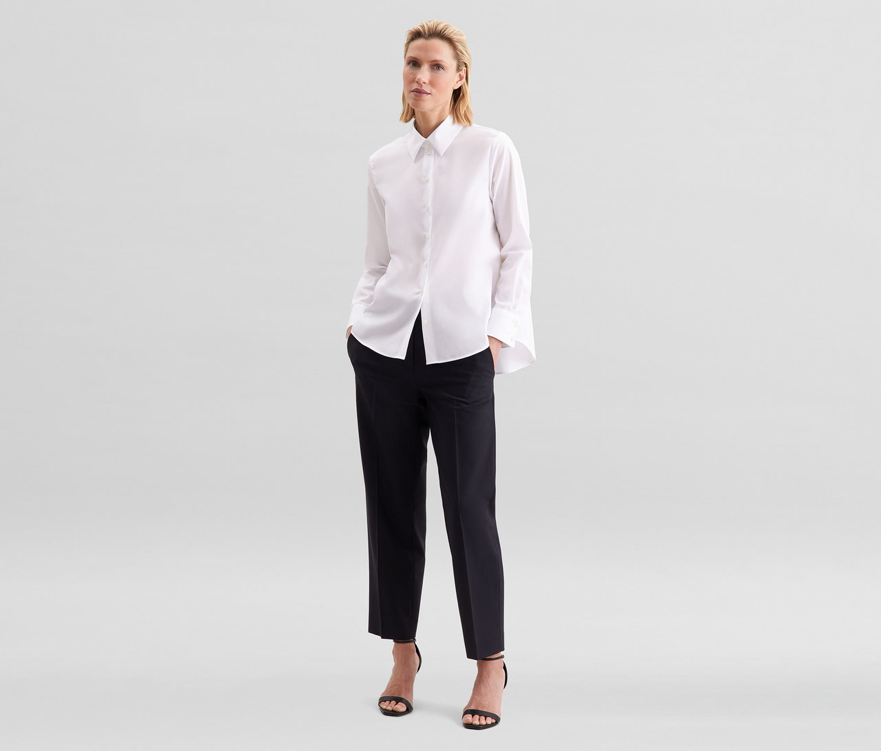 Blonde Frau steht, trägt eine weiße Seidensticker bügelfreie Bluse, schwarze Cropped-Hose und schwarze Sandalen mit hohen Absätzen.