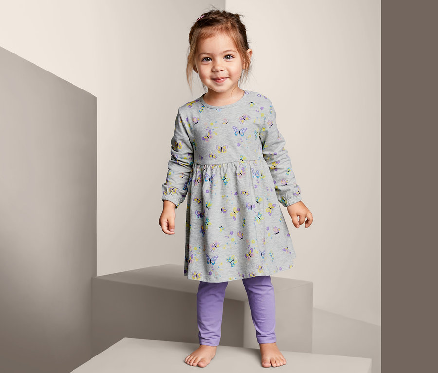 Ein lächelndes Mädchen steht auf einer Stufe und trägt ein graues Jerseykleid mit Leggings mit Schmetterlingsprint.