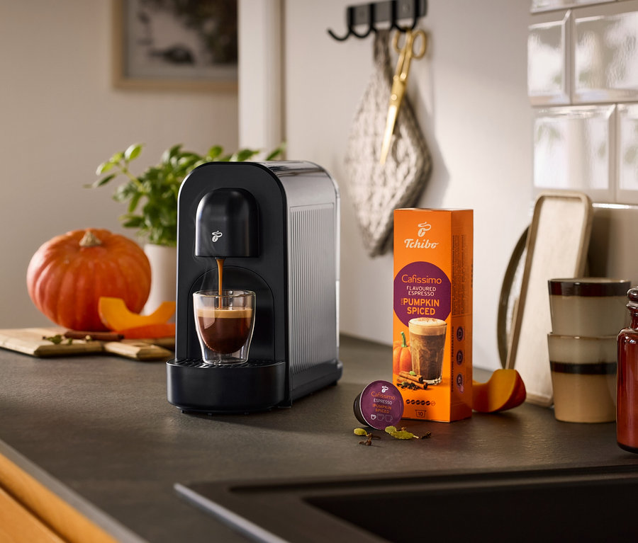Schwarze Cafissimo now Kaffeemaschine brüht Espresso in ein Glas, daneben eine Packung Cafissimo Flavoured Espresso Pumpkin Spiced und eine Kapsel, umgeben von Kürbis und Gewürzen auf einer Küchenzeile.
