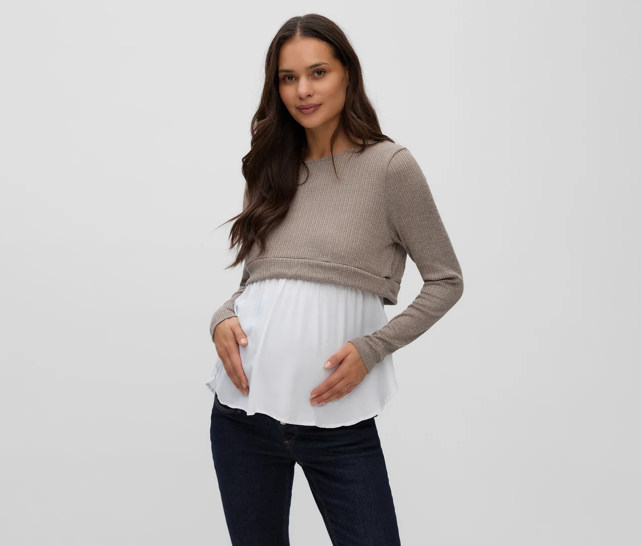 Eine schwangere Frau trägt ein MAMALICIOUS Mix-Top »MELONI JUNE« mit einem beigen gerippten Oberteil über einer weißen Lage und dunkelblauen Jeans. Sie hält sanft ihren schwangeren Bauch.