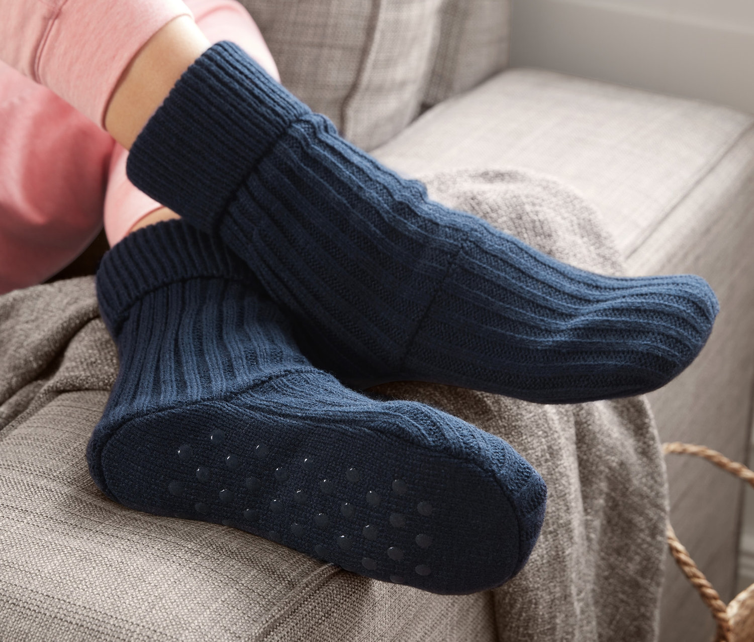 Beine einer Person in rosa Kleidung und marineblauen Hausschuhsocken sitzen auf einem grauen Sofa.